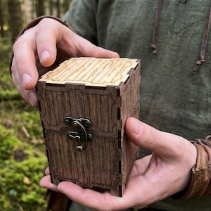 May include: A wooden box with a hinged lid and clasp, held in hands. The box has a textured, wood-grain appearance with decorative etched patterns. The lid and sides are a light brown colour, while the body is a darker brown.