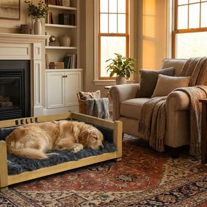 May include: A golden retriever dog sleeping in a wooden dog bed with a gray faux fur cushion. The bed is in a living room with a fireplace, armchair, and a side table with a lamp. The dog bed has the name "BELLE" on it.