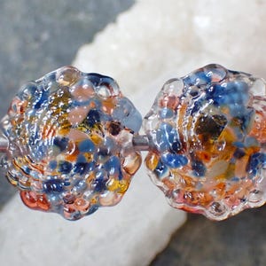 Op de afbeelding: Twee ambachtelijke glazen kralen met een getextureerd, bubbelachtig oppervlak. De kralen zijn veelkleurig, met tinten blauw, oranje, geel en helder glas. Ze zijn geregen op een zilverkleurige draad, perfect voor het maken van sieraden.