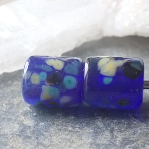 May include: Two square, cobalt blue glass beads with colorful inclusions of yellow, teal, and black. The beads are strung on a silver-colored metal rod, and are set against a neutral background.