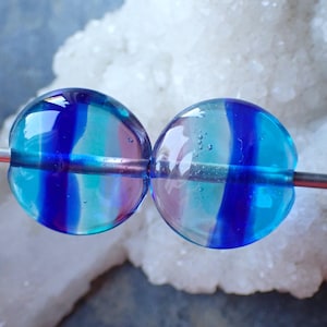 Peut inclure: Deux perles rondes en verre, rayées de bleu, turquoise et violet, sont enfilées sur un fil d'argent. Les perles transparentes laissent passer la lumière. Le fond est une structure cristalline blanche.