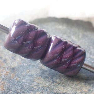 May include: Two dark purple glass beads with a twisted rope design are strung on a silver-colored metal rod. The beads have a glossy finish and are displayed against a natural stone background.
