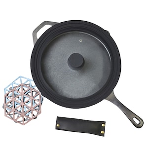 May include: A black cast iron skillet with a silicone lid and a black leather handle. The skillet is on a white background. There are also three geometric trivet coasters in blue, pink, and brown.