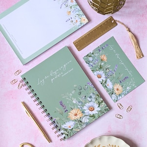 Pode incluir: Um caderno espiral verde com o texto "Lay me down in green pastures" e ilustrações florais. O caderno está sobre uma superfície rosa com uma régua dourada, um marcador dourado e um objeto decorativo dourado.
