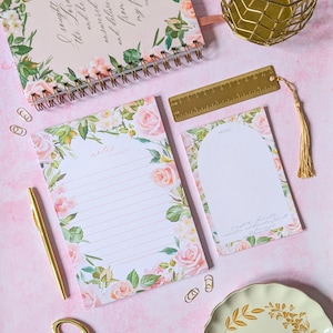 May include: Two floral patterned notepads with pink flowers and green leaves. The top notepad has a gold spiral binding and the text "I sought the Lore and he answered and delivered me from my f" printed on the cover. The bottom notepad has the text "notes & moments" printed on the cover.