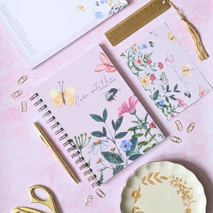 Op de afbeelding: Een roze spiraalgebonden notitieboek met een bloemenontwerp en de tekst "Come unto Me Devotional Journal". Het notitieboek is omringd door gouden paperclips, een gouden liniaal, een gouden kwast en een gouden pen.