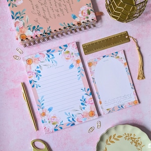 May include: Stationery set with a floral design, including a notebook, notepads, and a gold pen. The items feature a pink and white floral design with blue accents. A gold metal ruler and paperclips are also present.