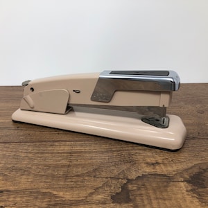 May include: A beige metal stapler with a silver top and a black plastic handle. The stapler is on a wooden surface.