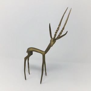 May include: A gold-colored metal sculpture of an antelope with long, twisted horns. The sculpture is stylized and abstract, with a simple, geometric design.