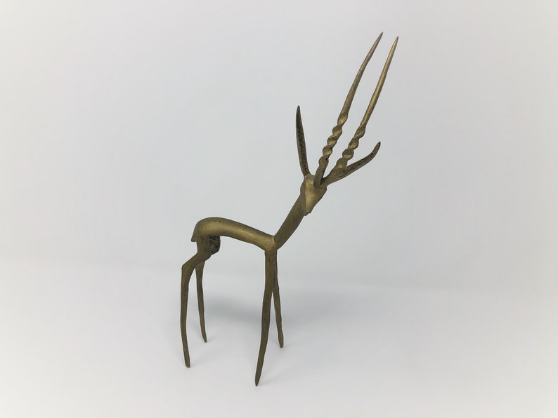 May include: A gold-colored metal sculpture of an antelope with long, twisted horns. The sculpture is stylized and abstract, with a simple, geometric design.