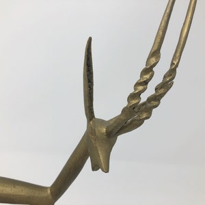 May include: A gold-colored metal sculpture of an antelope with twisted horns. The sculpture is a stylized representation of the animal, with a simplified body and long, thin legs.