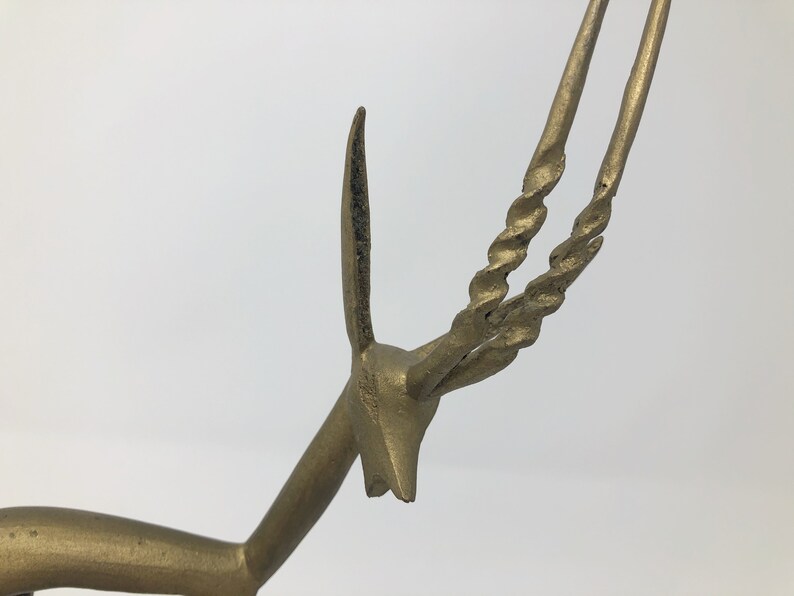 May include: A gold-colored metal sculpture of an antelope with twisted horns. The sculpture is a stylized representation of the animal, with a simplified body and long, thin legs.