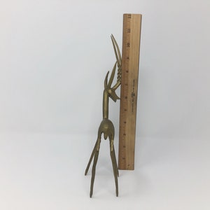 May include: A gold-colored metal sculpture of a stylized deer with long, slender legs and a raised head. The sculpture is about 9 inches tall and is standing on a white surface.
