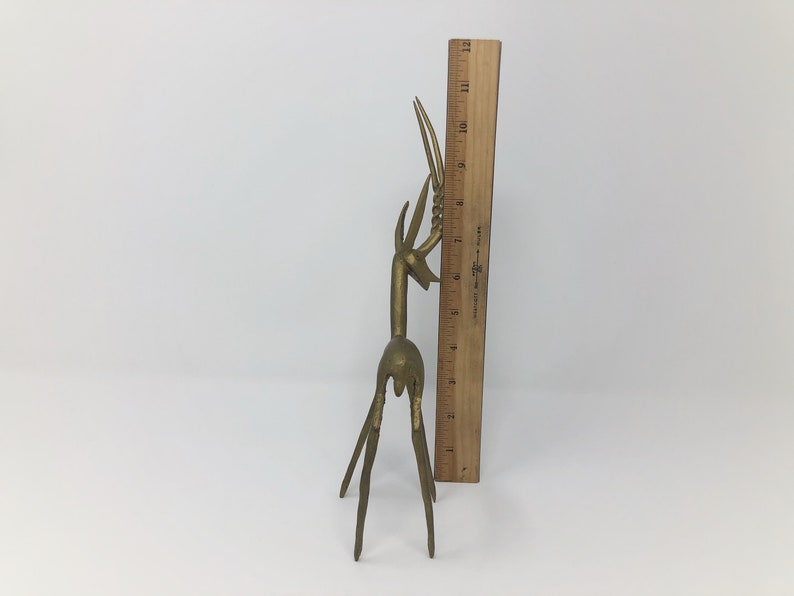 May include: A gold-colored metal sculpture of a stylized deer with long, slender legs and a raised head. The sculpture is about 9 inches tall and is standing on a white surface.