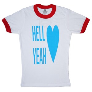 May include: White t-shirt with a red collar and sleeve trim. The shirt features the words "HELL YEAH" in light blue, along with a large blue heart graphic. The shirt has a classic ringer style.