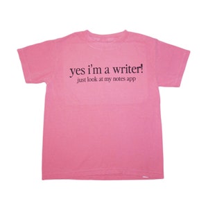 以下が含まれることがあります： ピンク色のTシャツ。黒字で「yes I'm a writer! just look at my notes app」と書かれています。Tシャツは丸首で半袖です。テキストは前面の中央に配置されています。