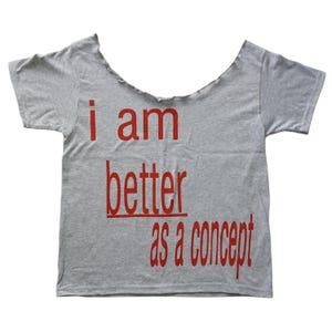 Puede incluir: Una camiseta gris con un cuello ancho y sin rematar y mangas cortas. La camiseta presenta el texto rojo "i am better as a concept" en negrita y sin serifa.