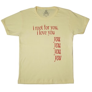 May include: A pale yellow t-shirt featuring the red text "i root for you, i love you". Beneath, the word "you" is repeated four times in red. The shirt has a crew neck and short sleeves. The brand name "SELF FIXATED" is printed at the top.
