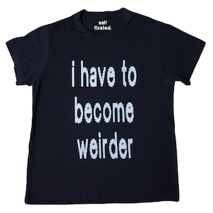 May include: A navy blue t-shirt with the text "i have to become weirder" in white block letters. The top of the shirt has the text "self fixated." in white. The shirt is a casual wear item.