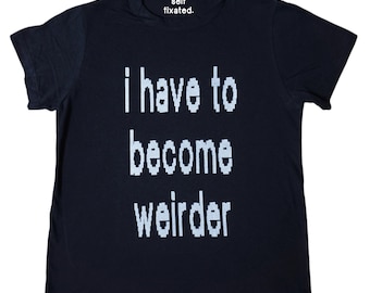 May include: A navy blue t-shirt with the text "i have to become weirder" in white block letters. The top of the shirt has the text "self fixated." in white. The shirt is a casual wear item.