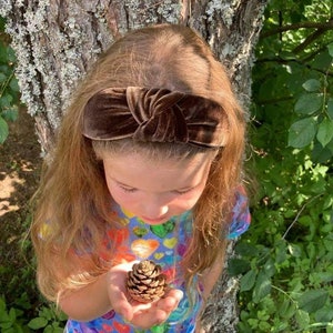 Può includere: Una bambina con lunghi capelli castani che indossa una fascia in velluto marrone e una camicia colorata con cuori stampati. Tiene un piccolo cono di pino marrone in mano.