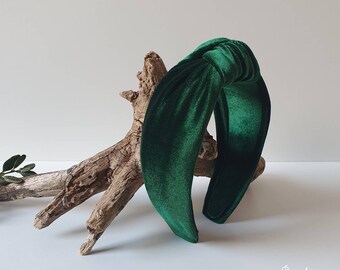May include: A dark green velvet headband with a knotted bow detail. The headband is displayed on a piece of driftwood.