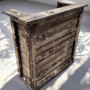 May include: A rustic wooden bar counter with a dark brown stain and a light wood grain. The counter has a simple design with a rectangular top and a square base.