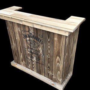 Op de afbeelding: Een houten bartafel met een Jack Daniel's logo gedrukt op de voorkant. De tafel is gemaakt van gerecycled hout en heeft een rustieke afwerking.