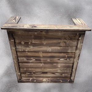 May include: A rustic wooden bar counter with a dark brown stain and a natural wood grain. The counter has a simple design with a flat top and a rectangular base.