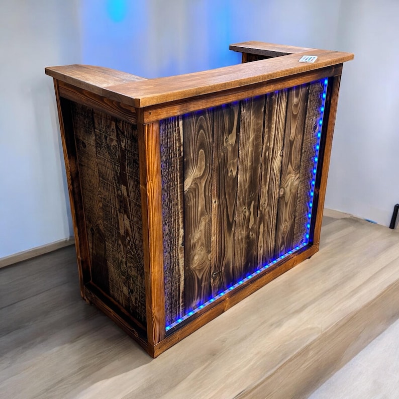 Op de afbeelding: Een rustieke houten bartafel met een donkerbruine afwerking en een blauwe LED-lichtstrip rond de onderkant. De tafel heeft een rechthoekig blad met een gebogen voorrand.