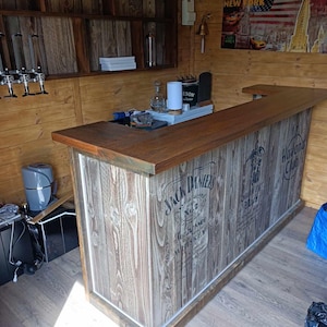 Op de afbeelding: Een houten bar met een bruin werkblad en een witte en grijze houtnerf afwerking. De bar is versierd met de logo's van Jack Daniel's en Havana Club.