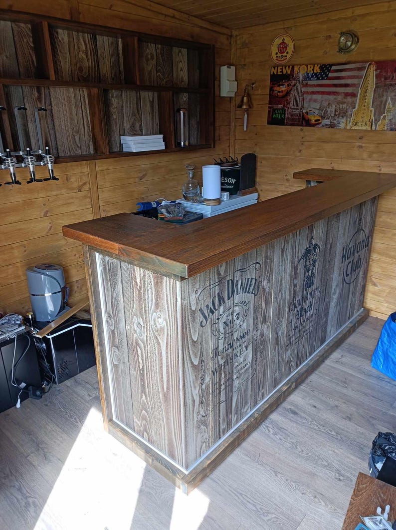 Op de afbeelding: Een houten bar met een bruin werkblad en een witte en grijze houtnerf afwerking. De bar is versierd met de logo's van Jack Daniel's en Havana Club.