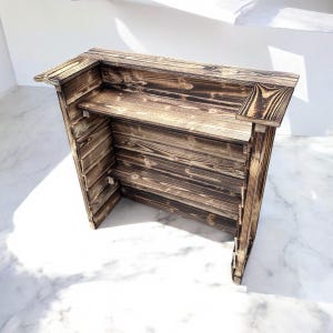 May include: A rustic wooden bar counter with a distressed finish. The counter has a shelf and a raised top. The wood is a dark brown with lighter streaks.