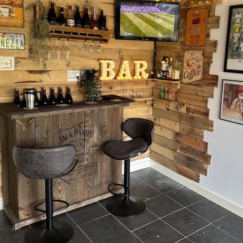 Op de afbeelding: Een houten bar met een Jack Daniels logo, twee zwarte barstoelen en een bord met de tekst "BAR" in lichten. De bar is versierd met houten lambrisering en verschillende borden, waaronder een met de tekst "Steak House" en een ander met de tekst "Coffee".