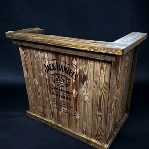 Op de afbeelding: Een rustieke houten bartafel met een Jack Daniel's logo gedrukt op de voorkant. De tafel is gemaakt van gerecycled hout en heeft een natuurlijke bruine afwerking.