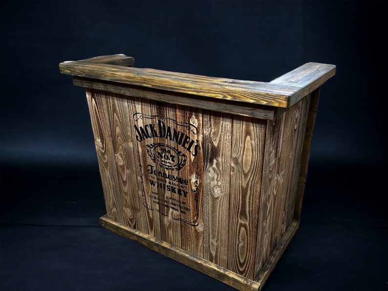 Op de afbeelding: Een rustieke houten bartafel met een Jack Daniel's logo gedrukt op de voorkant. De tafel is gemaakt van gerecycled hout en heeft een natuurlijke bruine afwerking.