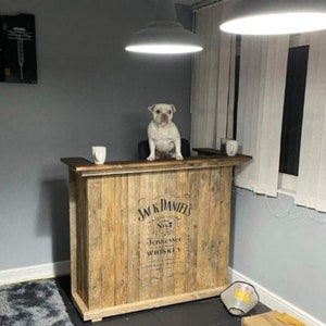 Op de afbeelding: Een houten bartafel met een Jack Daniel's Tennessee Whiskey logo. De tafel heeft een lichte houtkleur en een witte hond zit erop.
