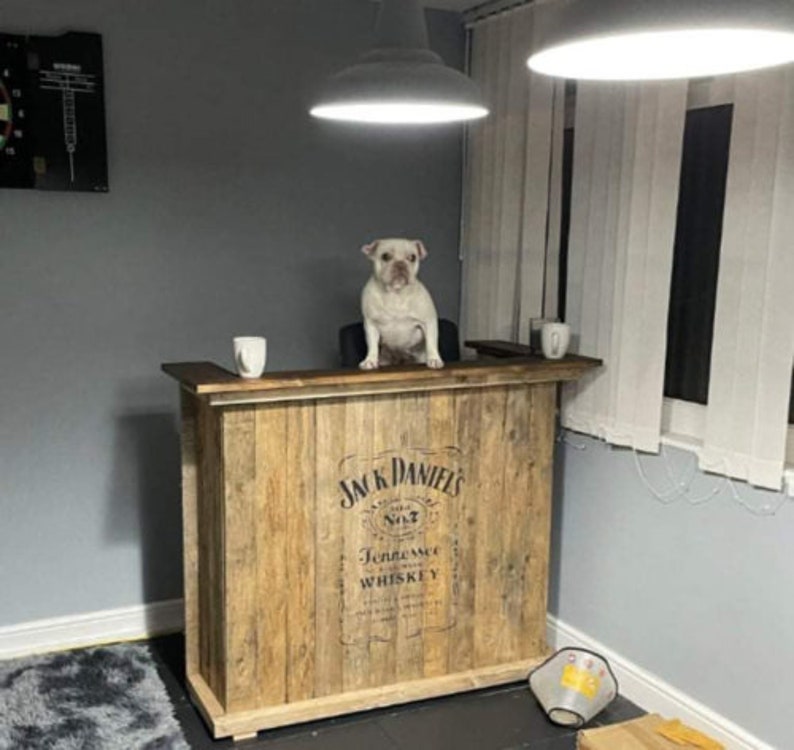 Op de afbeelding: Een houten bartafel met een Jack Daniel's Tennessee Whiskey logo. De tafel heeft een lichte houtkleur en een witte hond zit erop.