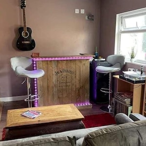 Op de afbeelding: Een houten bar met een Jack Daniel's whiskey-label, paarse LED-verlichting en twee witte barstoelen. De bar bevindt zich in een kamer met een gitaar aan de muur, een platenspeler en een boekenkast vol platen.