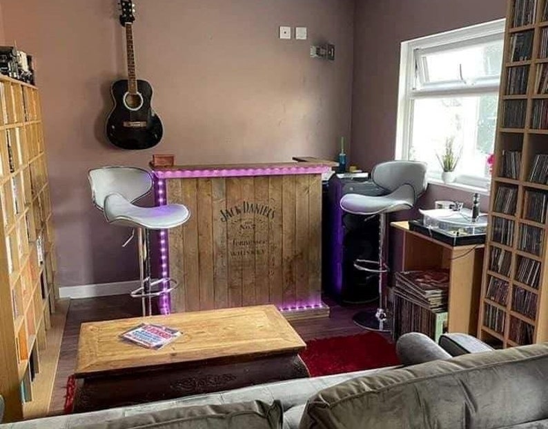 Op de afbeelding: Een houten bar met een Jack Daniel's whiskey-label, paarse LED-verlichting en twee witte barstoelen. De bar bevindt zich in een kamer met een gitaar aan de muur, een platenspeler en een boekenkast vol platen.