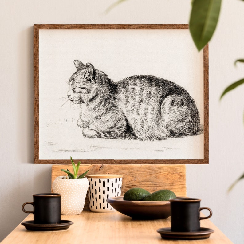 May include: A black and white drawing of a cat sitting in a wooden frame. The frame is hanging on a white wall. There are two coffee mugs and a potted plant on a wooden table in the foreground.