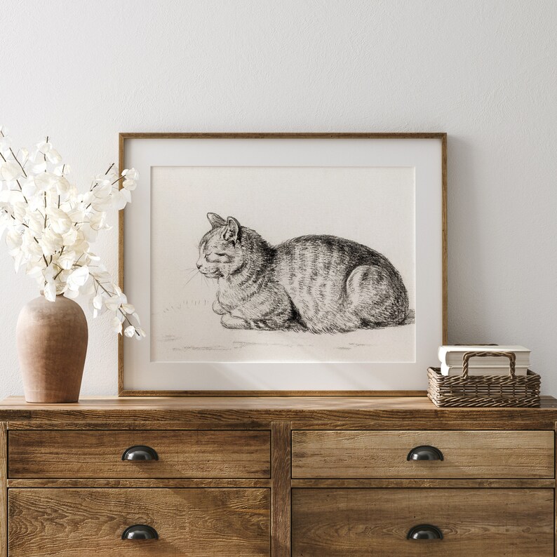 May include: A black and white line drawing of a cat curled up in a sleeping position. The drawing is framed in a light brown wood frame and is hanging on a white wall.