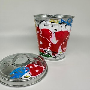 May include: A silver metal trash can with a lid. The can is painted with red, white, blue, yellow, and black graffiti. The graffiti includes the word "B.Y.B."