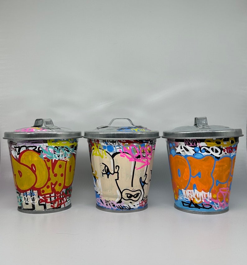 May include: Three silver trash cans with colorful graffiti designs. The cans are painted with various colors, including yellow, blue, orange, pink, and white. The designs include letters, shapes, and abstract patterns.