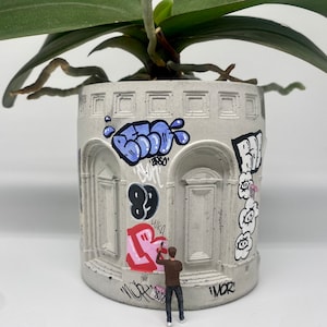 Puede incluir: Una maceta de hormigón gris con arte de graffiti, con una figura en miniatura pintando con aerosol el lateral. La maceta tiene un diseño arquitectónico con aberturas arqueadas. Una planta de orquídea con hojas verdes y raíces expuestas está encima.