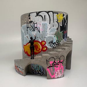 May include: A cylindrical concrete planter featuring a staircase wrapping around the exterior. The surface is covered in colorful graffiti tags and murals in black, white, red, yellow, and pink. A small black figurine stands on the stairs.