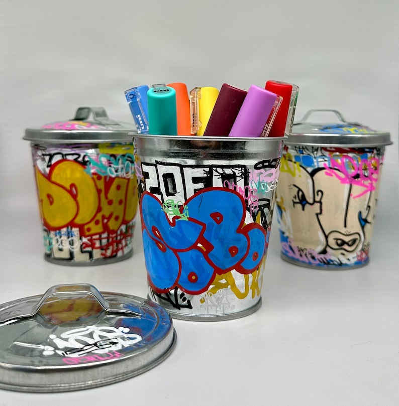 May include: Four small metal trash cans with graffiti designs. The cans are filled with colorful markers.