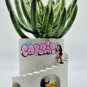 May include: A white concrete planter shaped like a spiral staircase with graffiti art, including the word "BARBIE" in pink letters. A small figurine of a person is painting the graffiti. The planter is filled with a green succulent plant.