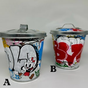May include: Two small metal trash cans with lids, painted with colorful graffiti designs. One can features a cartoon character, while the other has a red and white graffiti design. The cans are labeled A and B.