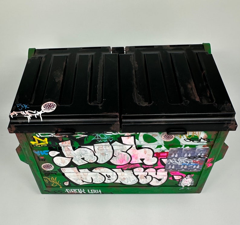 May include: A green metal dumpster with a black lid. The dumpster is covered in colorful graffiti, including the words "Junk" and "Love".